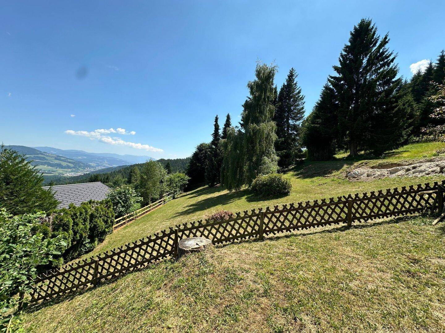 Sonnige Rasenfläche am Hang mit rustikalem Holzzaun und Panoramablick auf die bewaldeten Berge in der Ferne.