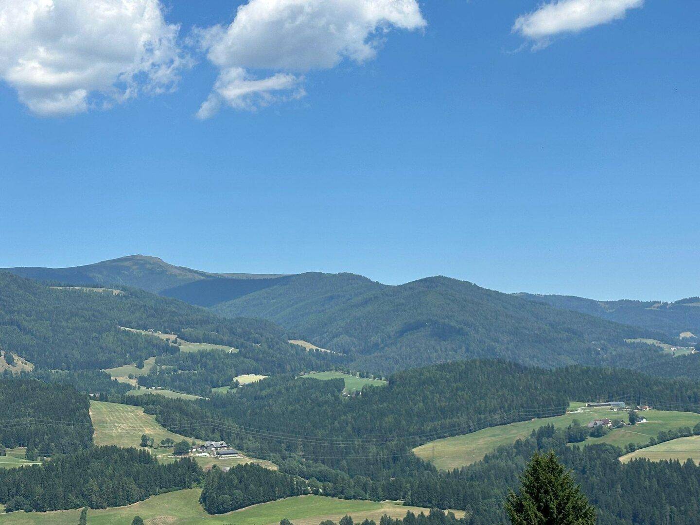 Weitläufiger Blick über die sanften Hügel und bewaldeten Täler der Berglandschaft unter blauem Himmel.
