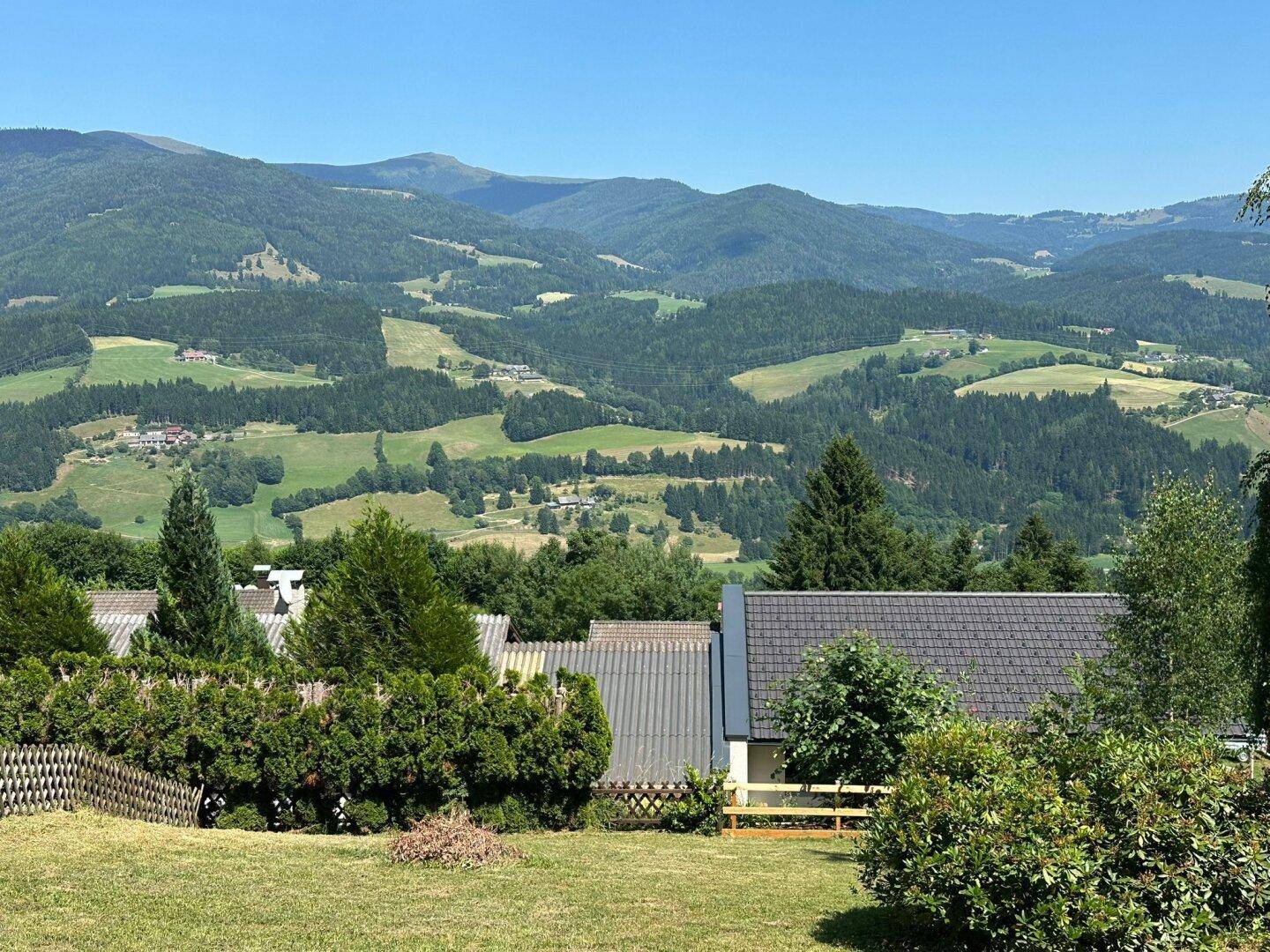 Blick auf die Dächer der Häuser und die dahinterliegende, bewaldete Bergkette an einem klaren Sommertag.