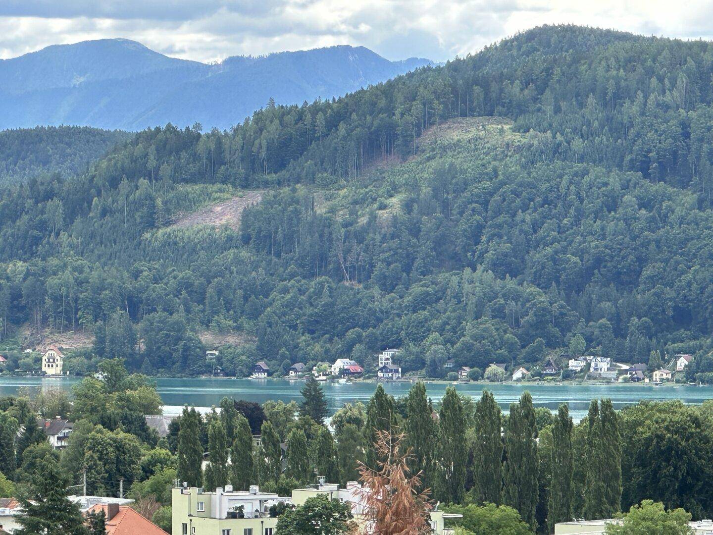 Panoramablick auf den Wörthersee mit Häusern am Ufer und bewaldeten Bergen im Hintergrund.