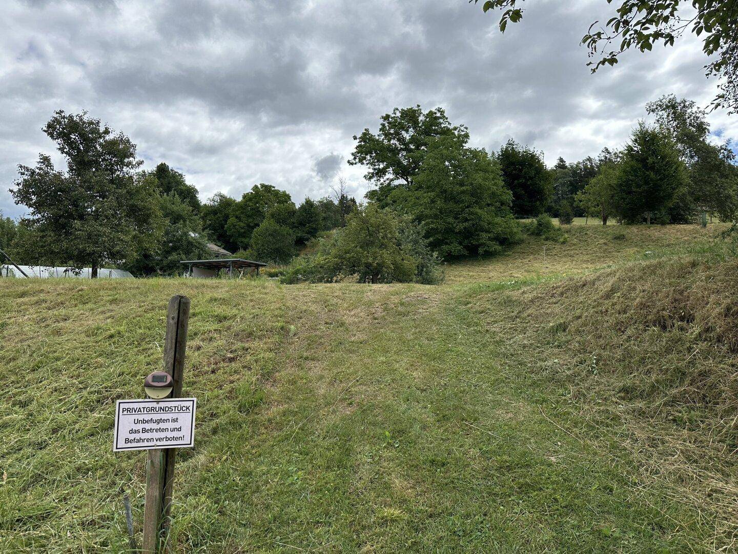 Eine abfallende Wiese mit einem Schild "Privatgrundstück" und Bäumen unter bewölktem Himmel.