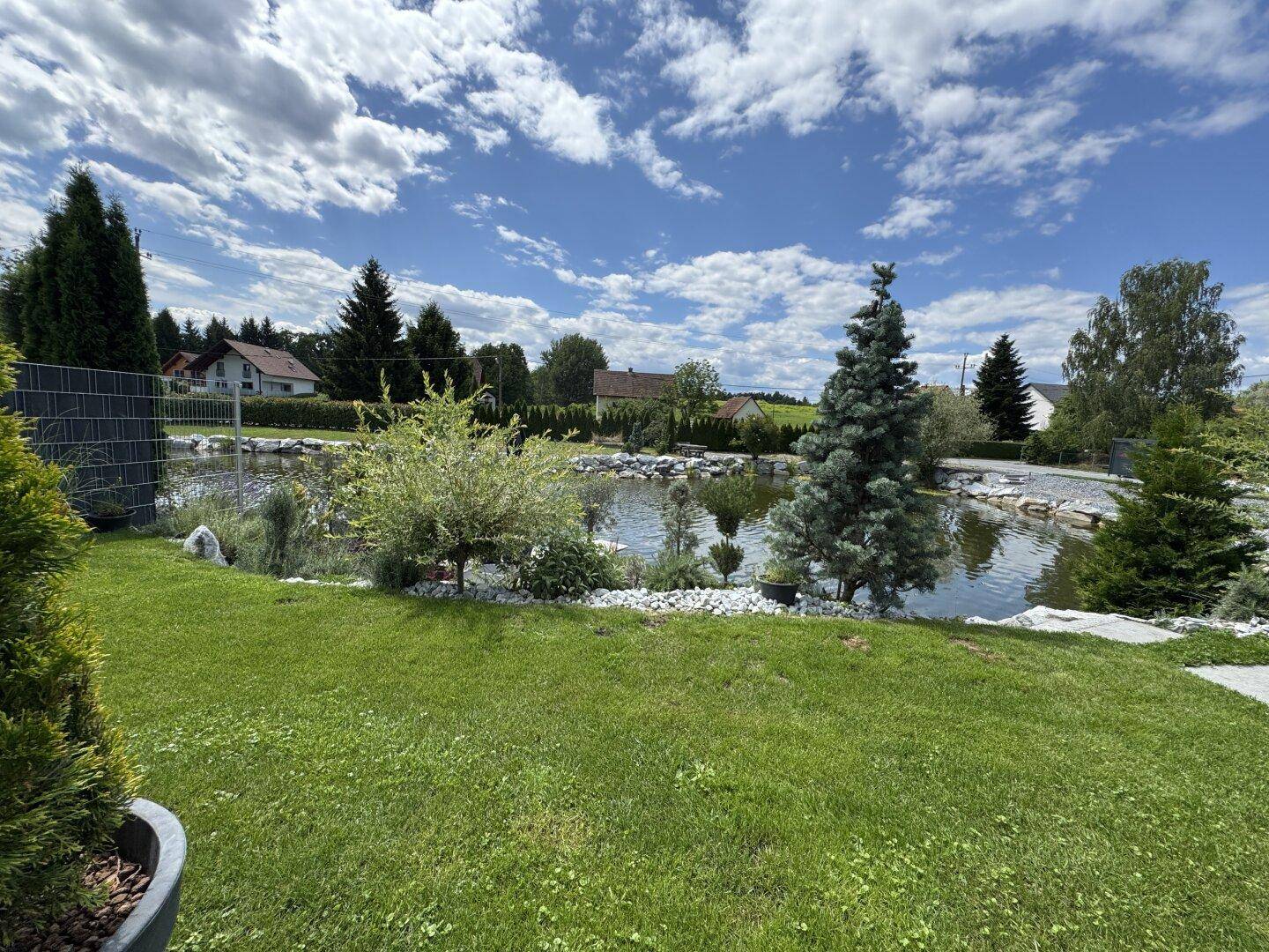 Weitläufiger Gartenbereich mit großem Naturteich, umgeben von Rasenflächen und Bäumen unter blauem Himmel.
