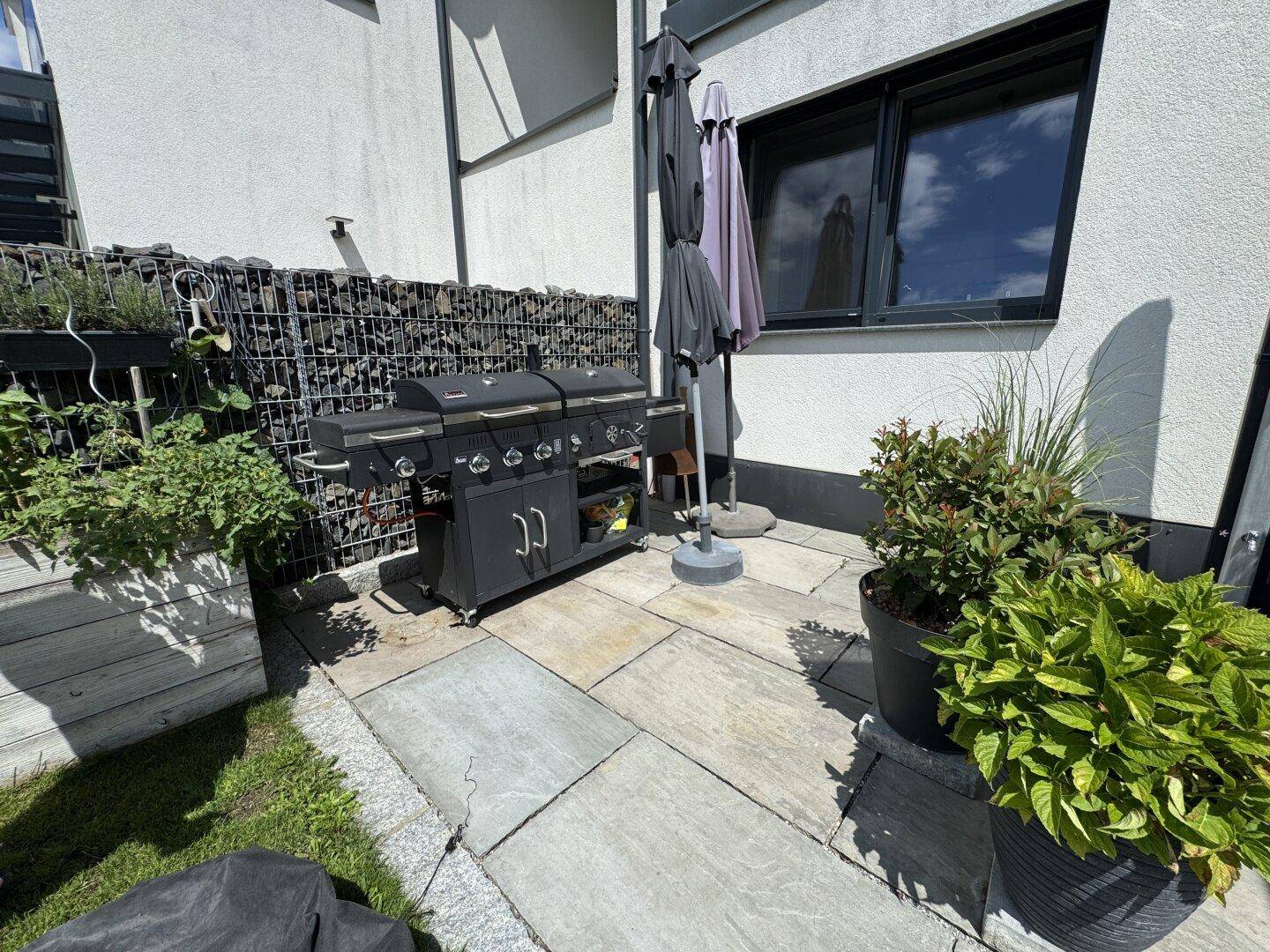 Gemütlicher Grillbereich auf einer gepflasterten Terrasse mit großem Gasgrill und Gabionenwand.