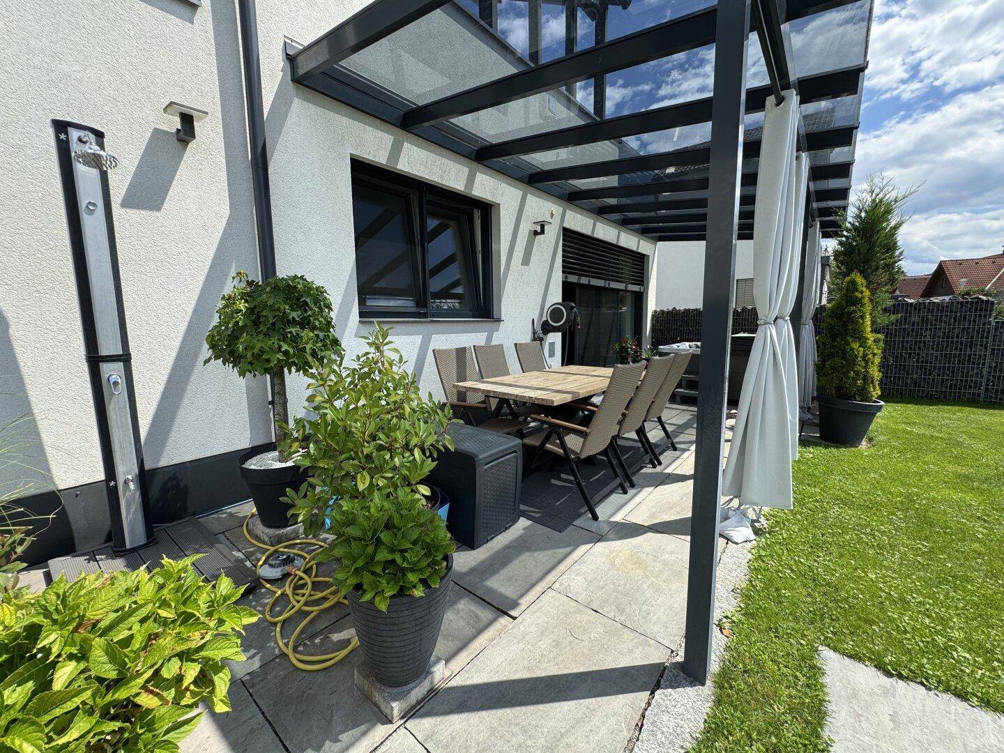 Überdachte Terrasse mit Esstisch, Stühlen und Außendusche, umgeben von Grünflächen.