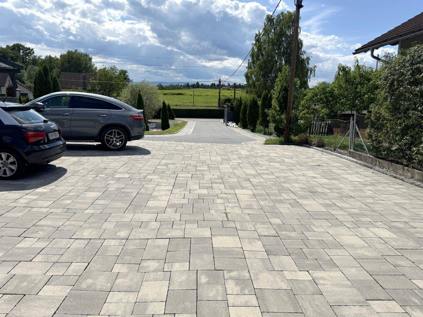 Gepflasterte Einfahrt mit Parkplätzen und Blick auf die weite Landschaft unter bewölktem Himmel.