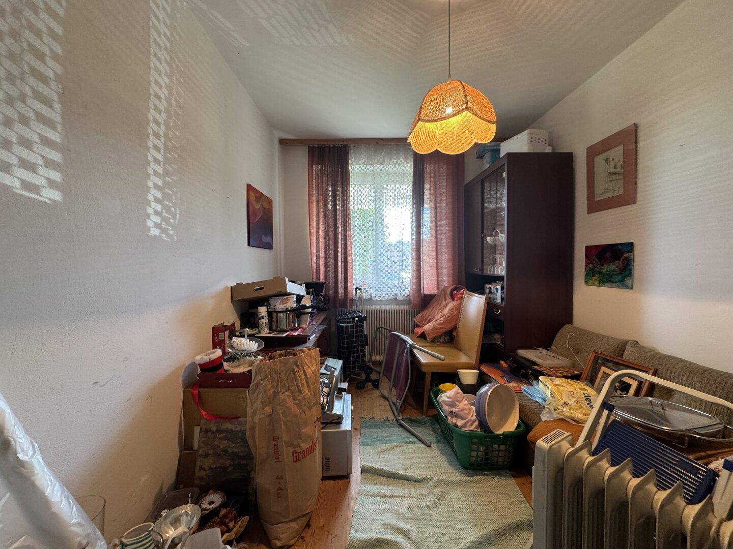 Unaufgeräumter Raum mit Fenster, Teppichboden und verschiedenen Gegenständen.