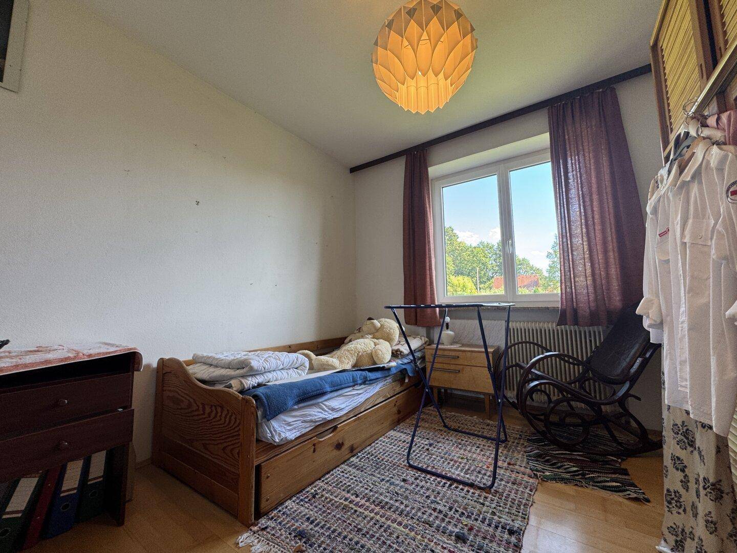 Schlafzimmer mit Einzelbett, Holzmöbeln, Teppich und Fenster mit Blick ins Grüne.