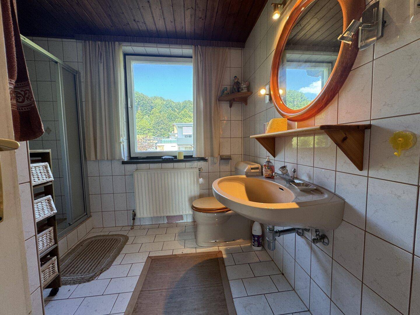 Badezimmer mit hellen Fliesen, Dusche, Waschbecken und Fenster mit Bergblick.