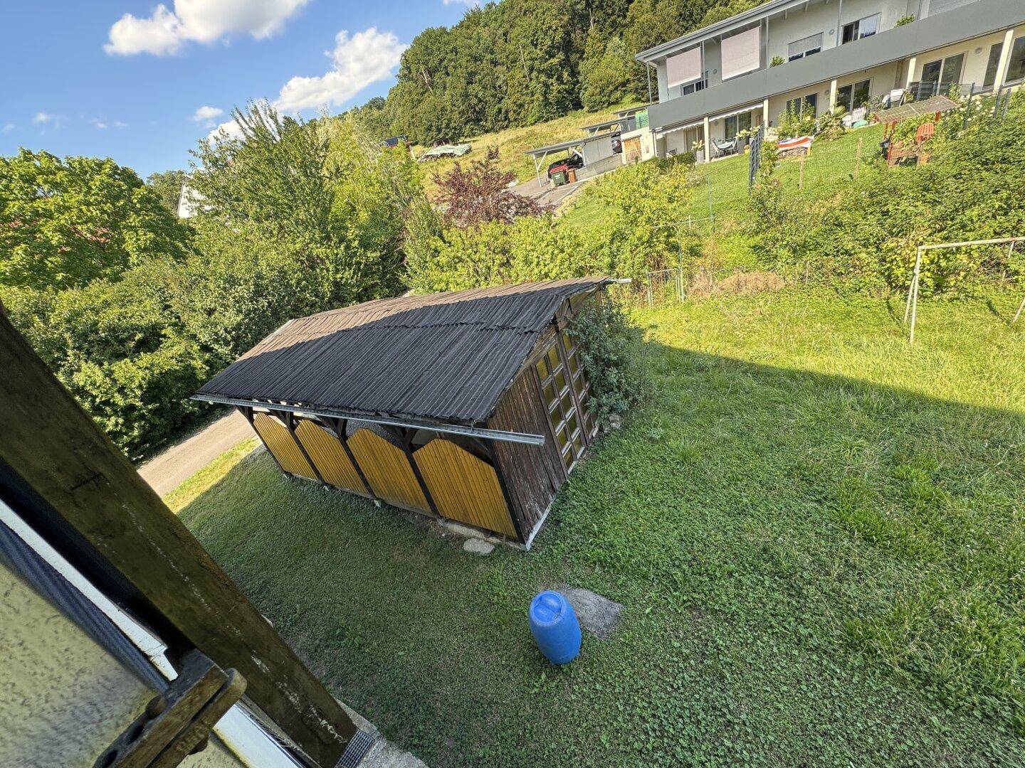 Kleiner Holzschuppen im Gartenbereich, umgeben von Grünflächen und Bäumen.