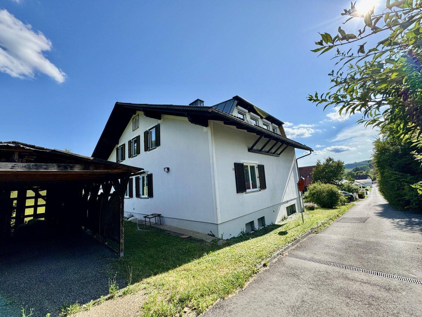 Seitenansicht des Hauses mit weißer Fassade, Carport und Zufahrt, umgeben von Grün.