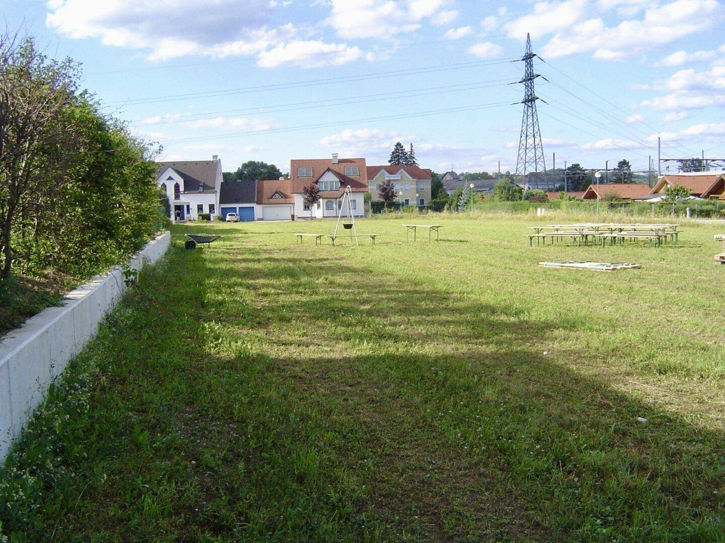 Weitläufige Rasenfläche mit Wohnhäusern im Hintergrund und einem Strommast am Horizont.