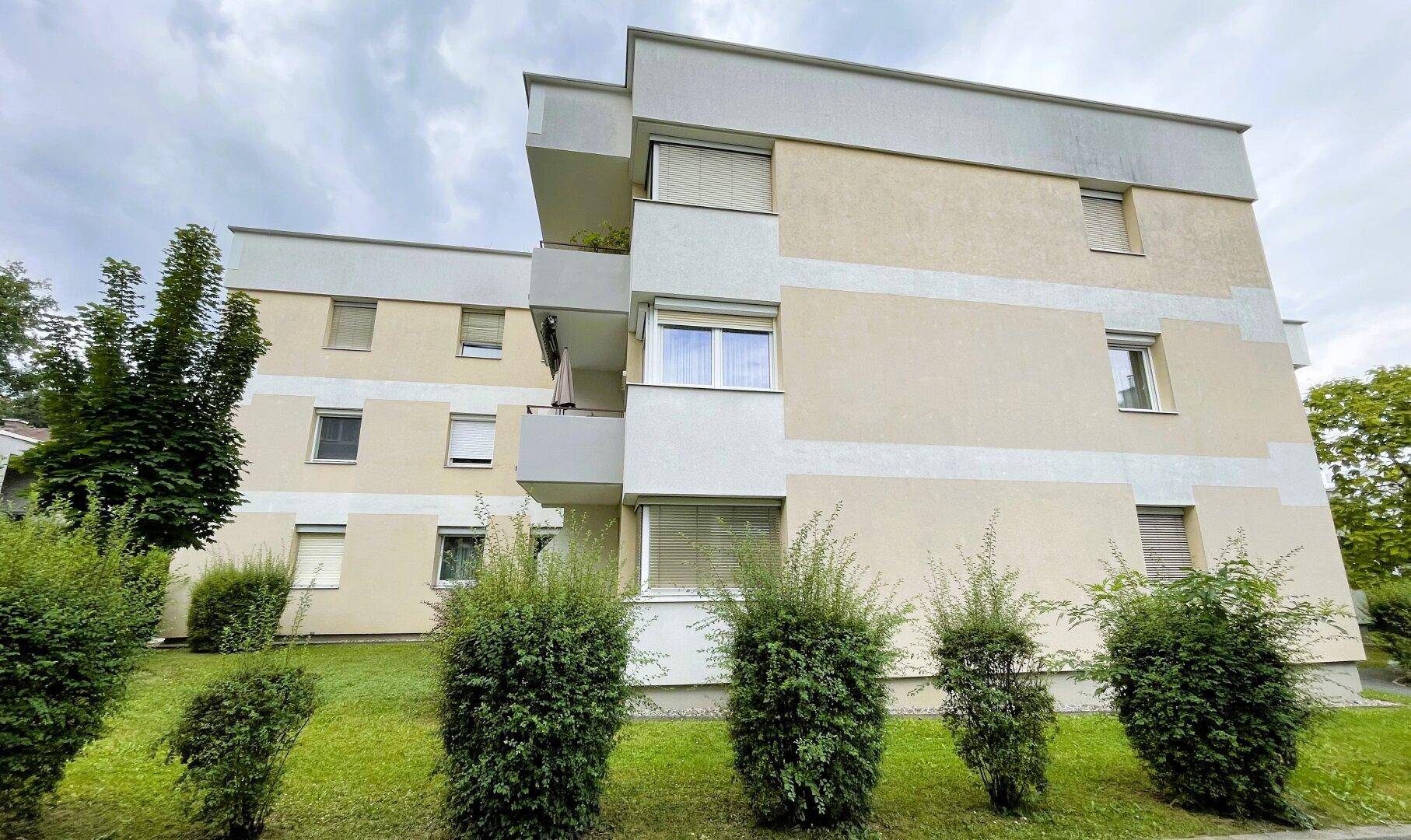 Mehrfamilienhaus mit heller Fassade, Balkonen und umliegender Grünfläche.