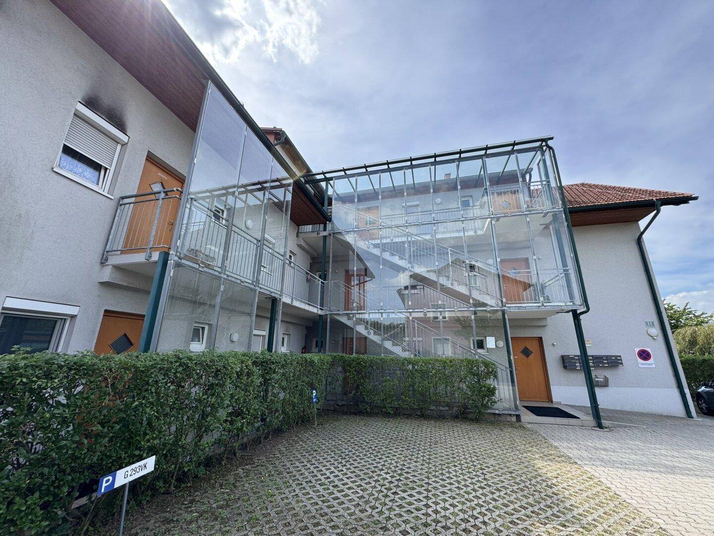 Außenansicht eines Wohngebäudes mit verglastem Treppenhaus und Parkplätzen vor einer Hecke.