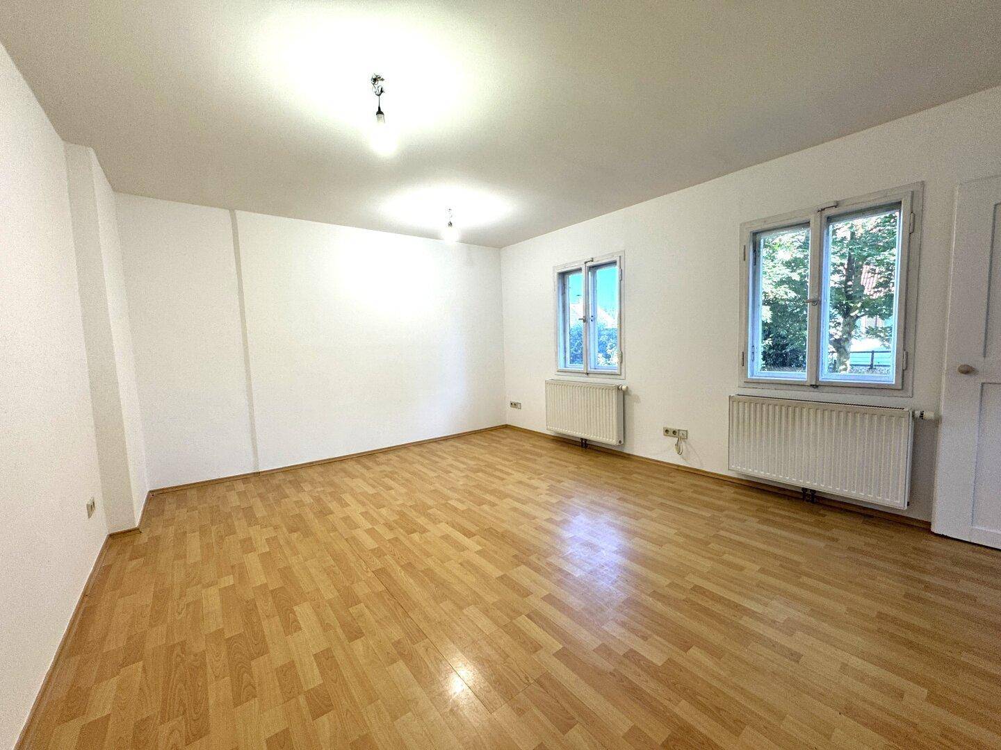 Geräumiges, helles Zimmer mit Holzparkett, weißen Wänden und Heizkörpern unter den Fenstern.