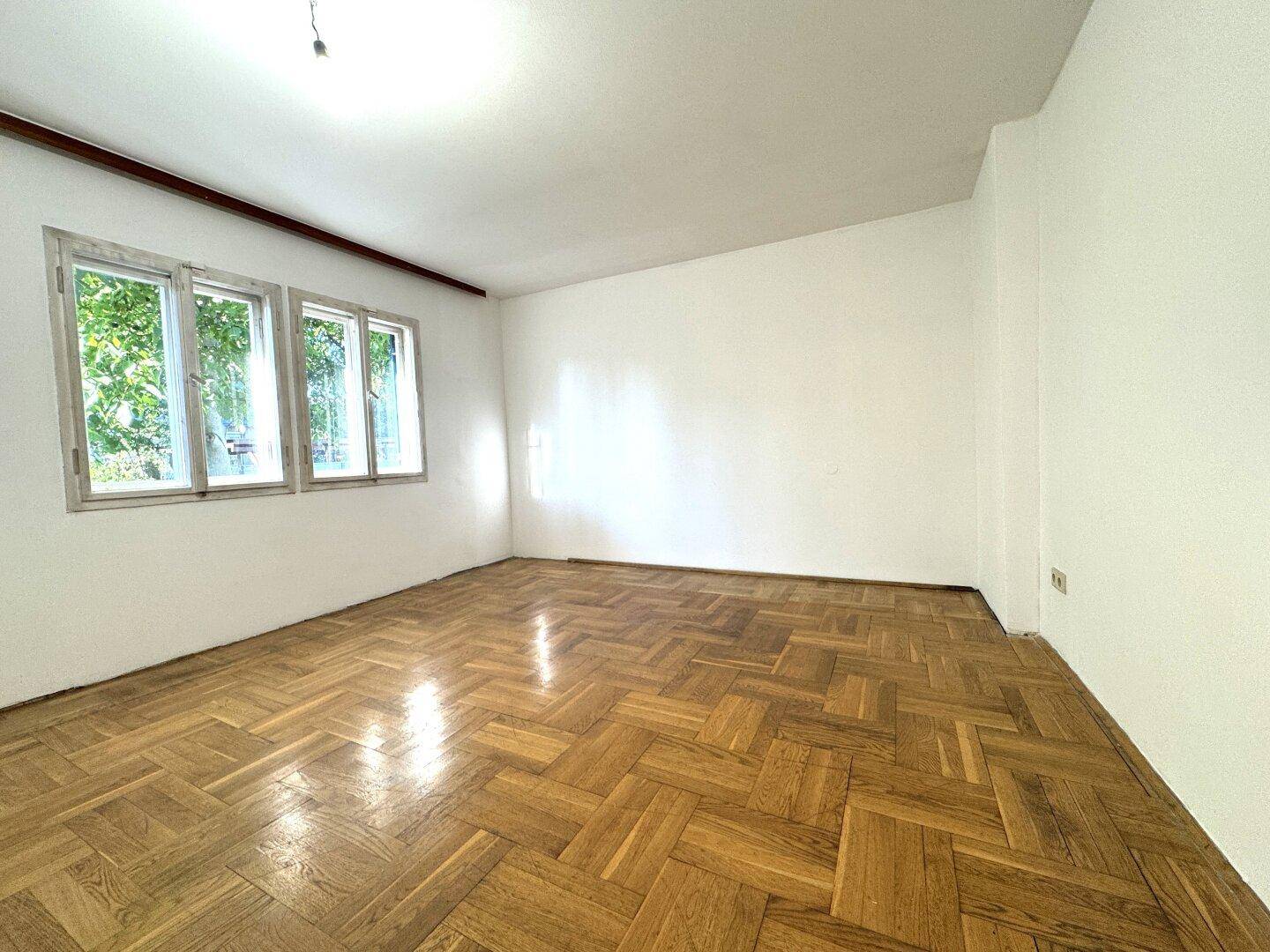Helles, leeres Zimmer mit Fischgrätparkettboden und großen Fenstern mit Blick ins Grüne.