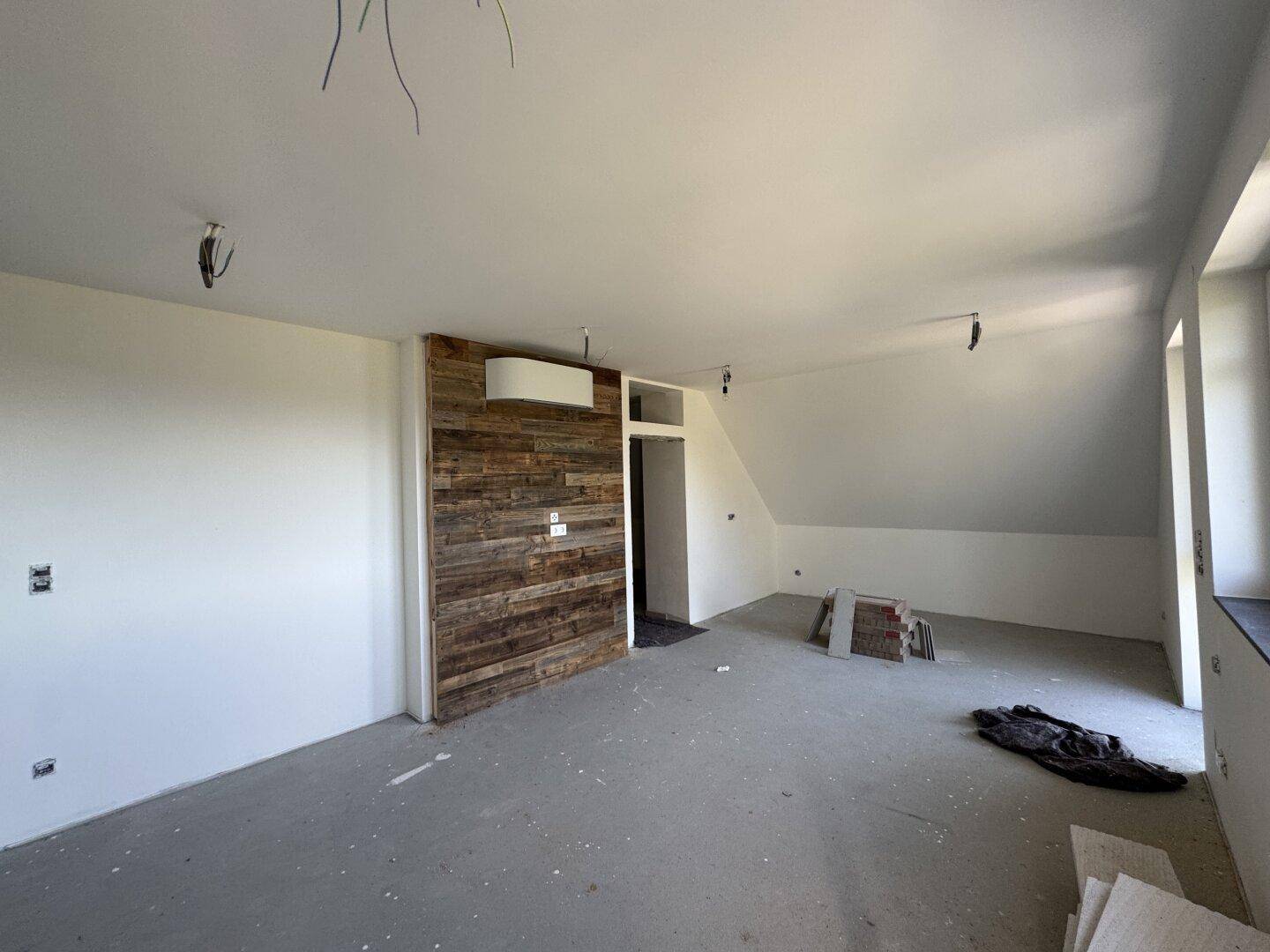Geräumiger Raum im Rohbau mit Holzakzentwand, schräger Decke und vorbereiteter Klimaanlage.
