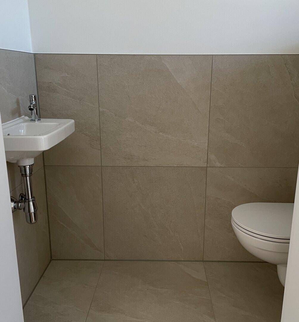 Modernes Gäste-WC mit beigefarbenen Fliesen, wandhängendem WC und kleinem Waschbecken.