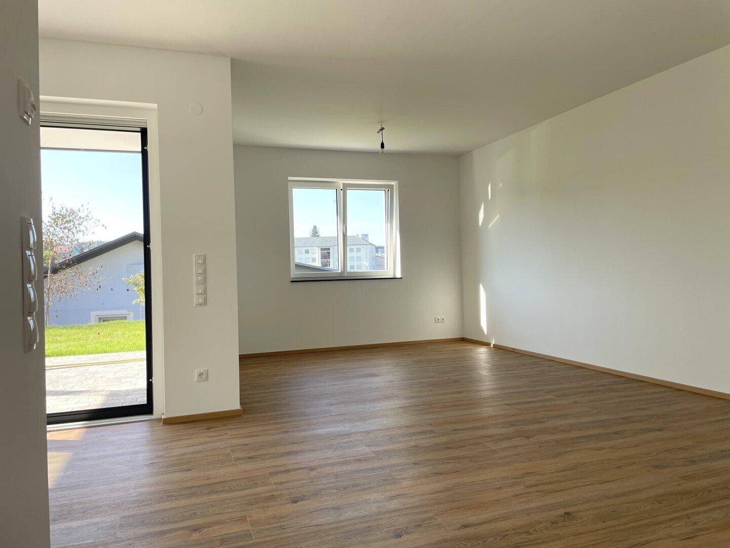 Geräumiger Wohnbereich mit Laminatboden und großen Fenstern, die viel Tageslicht hereinlassen.