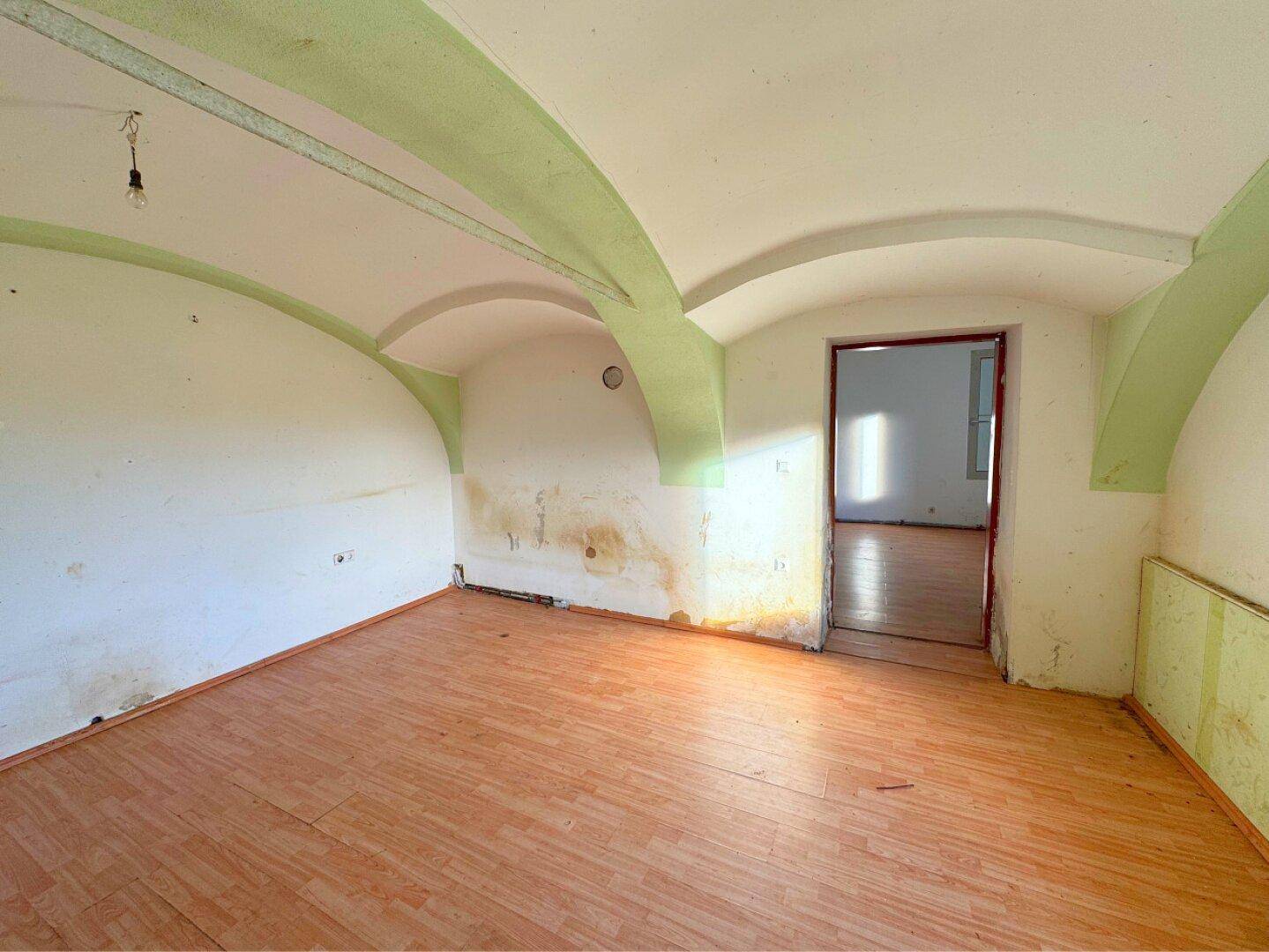Leerer Raum mit Gewölbedecke, Laminatboden und sichtbaren Feuchtigkeitsschäden, bereit zur Renovierung.