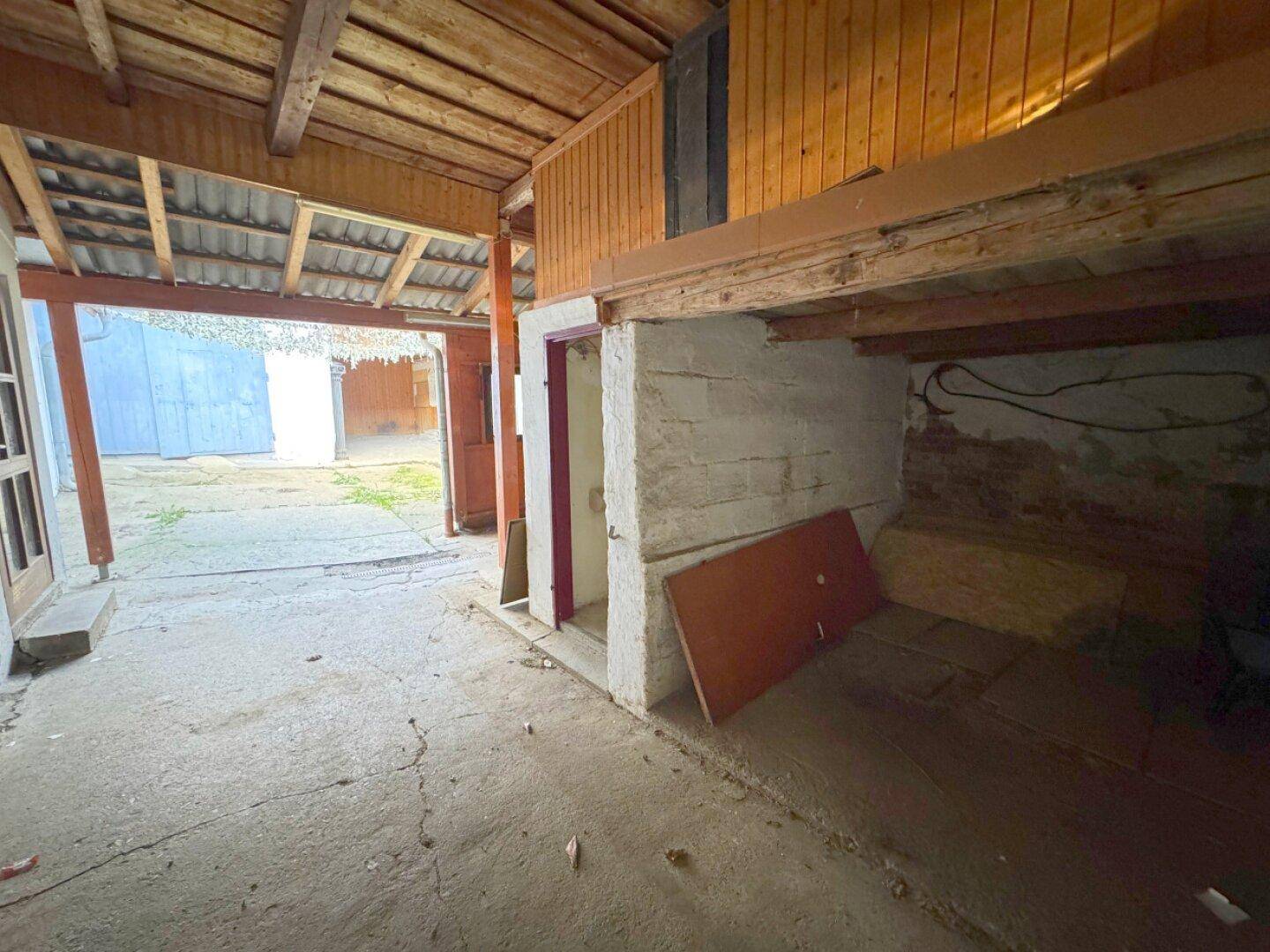 Rustikaler Innenraum eines Nebengebäudes mit Holzbalken, Betonboden und rohen Wänden, renovierungsbedürftig.