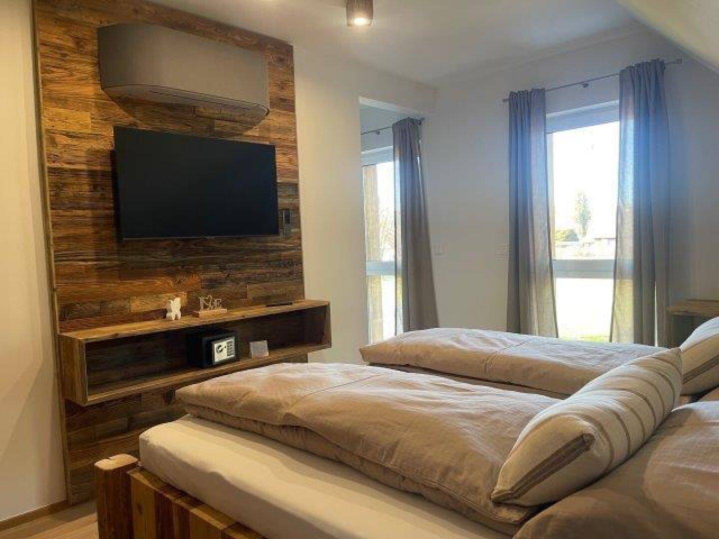 Schlafzimmer mit zwei Einzelbetten, einer Holzwand mit Fernseher und großem Fenster mit Vorhängen.
