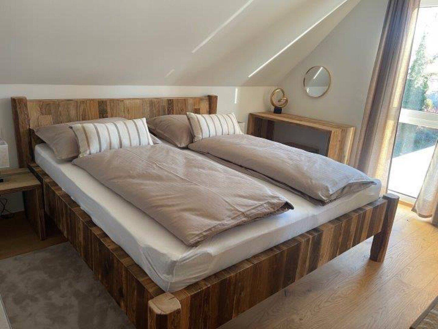 Helles Schlafzimmer mit rustikalem Holzbett, Holzboden und großem Fenster unter einer Dachschräge.