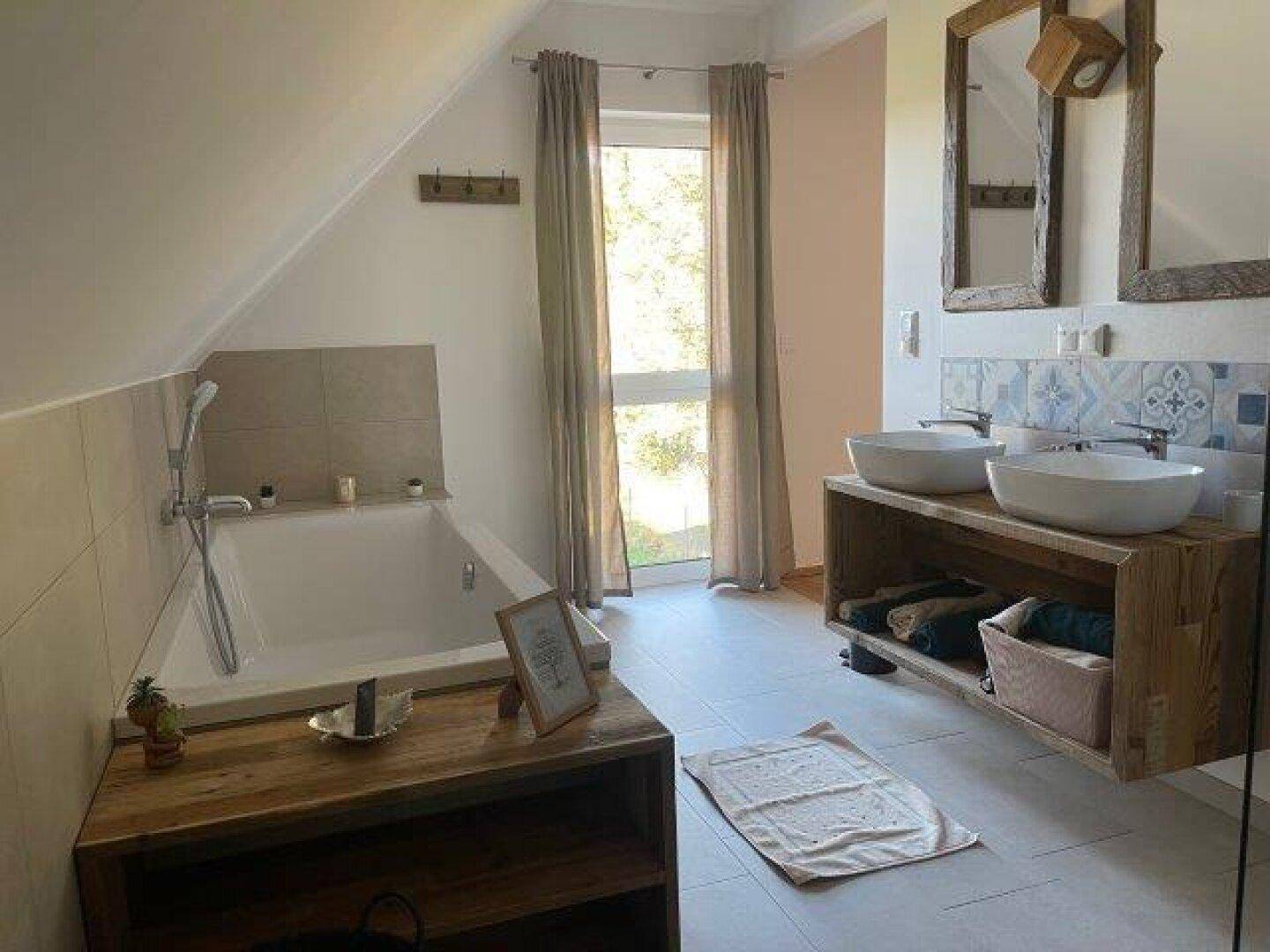 Helles Badezimmer mit Badewanne, Doppelwaschbecken auf Holzunterschrank und großem Fenster unter der Dachschräge.