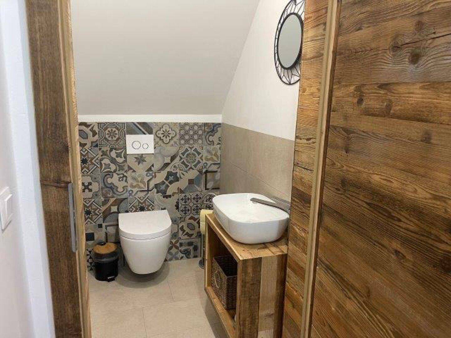 Gäste-WC mit Wand-WC, Einzelwaschbecken auf Holzunterschrank und gemusterten Fliesen unter der Dachschräge.