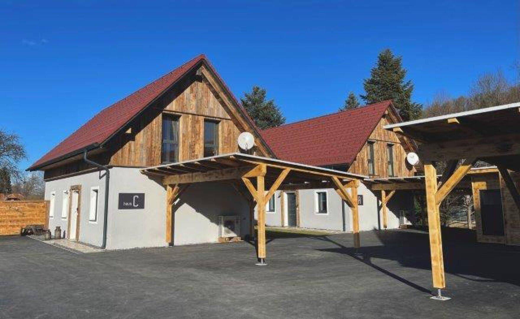 Zwei moderne Häuser mit roten Ziegeldächern und Holzfassaden, umgeben von Carports und gepflasterten Flächen.