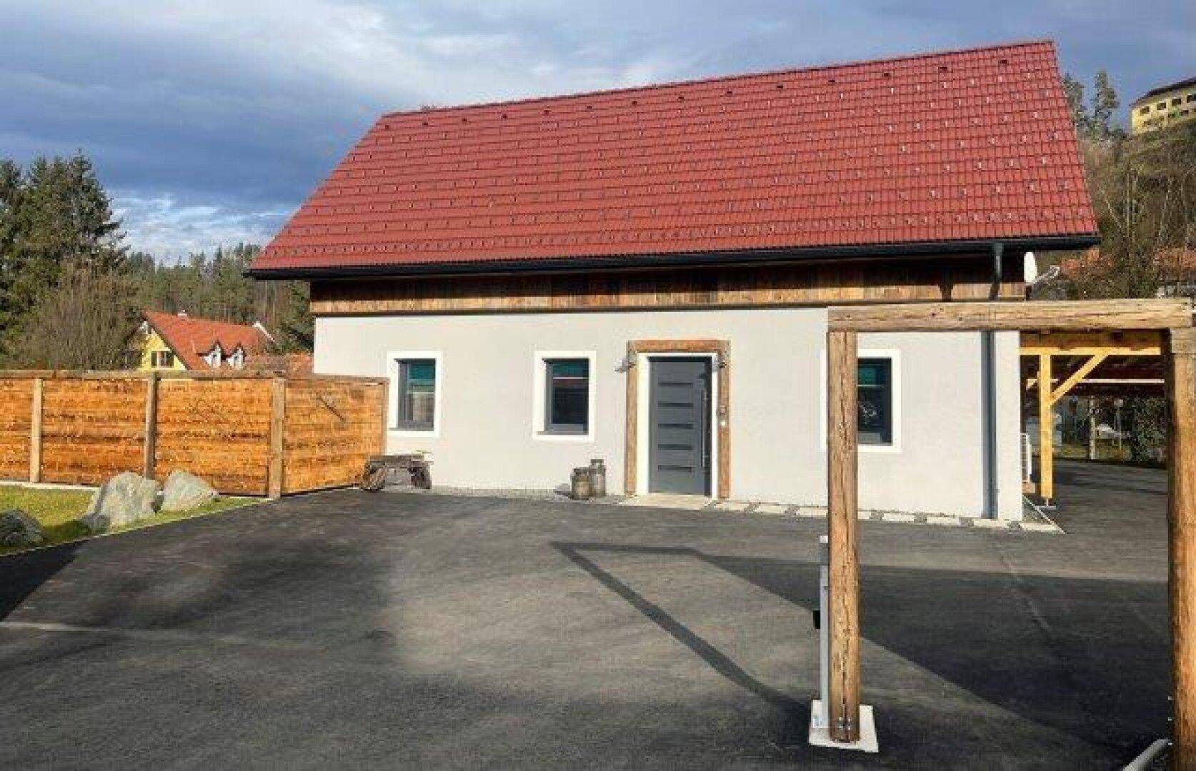 Ein modernes Haus mit heller Fassade, rotem Dach und einem hölzernen Carport auf einem gepflasterten Vorplatz.