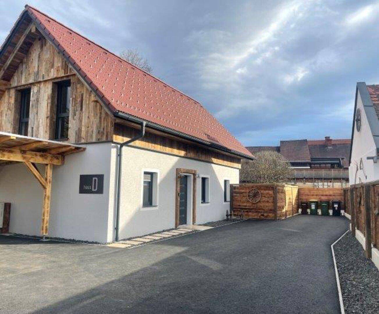 Modernes Haus mit heller Fassade, rotem Dach und Carport, umgeben von einem Holzzaun und gepflasterter Zufahrt.