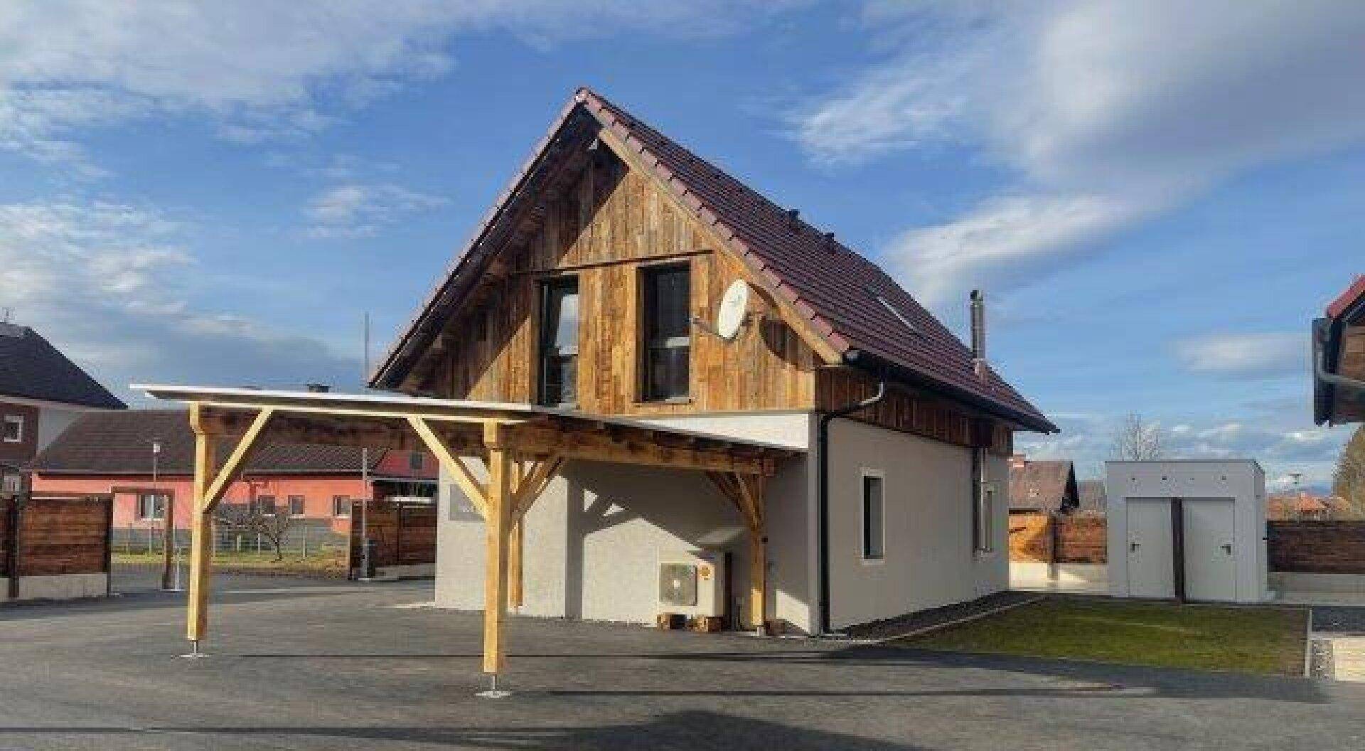 Modernes Haus mit Carport, kleiner Rasenfläche und einem weißen Abstellraum unter blauem Himmel.
