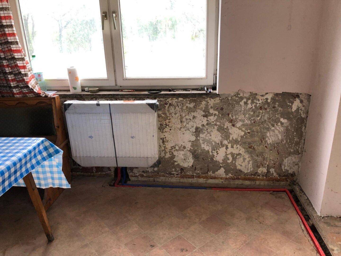 Küche in Renovierung mit Fenster, verpacktem Heizkörper und karierter Tischdecke.