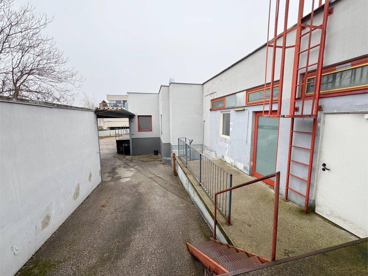 Zugangsbereich zum Gebäude mit Rampe und Treppe, umgeben von grauen Wänden.