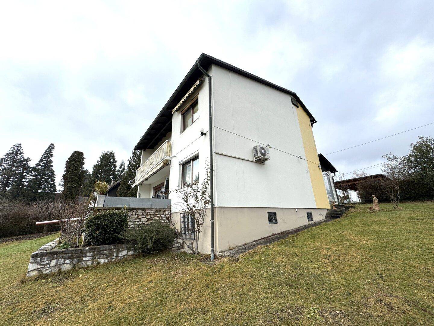 Seitenansicht des Hauses mit weißer Fassade, Rasenfläche und Bäumen im Hintergrund.