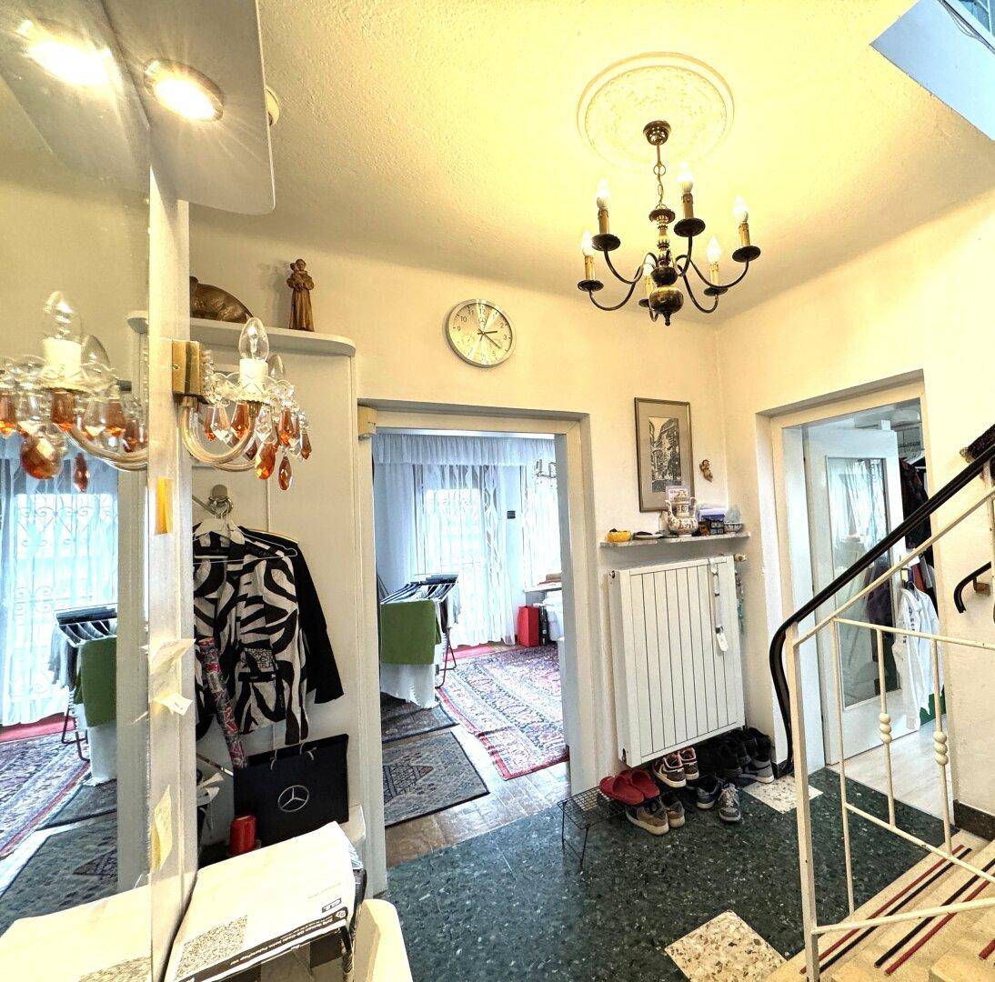 Eingangsbereich mit Treppe, Kronleuchter und Terrazzoboden, der zu weiteren Räumen führt.