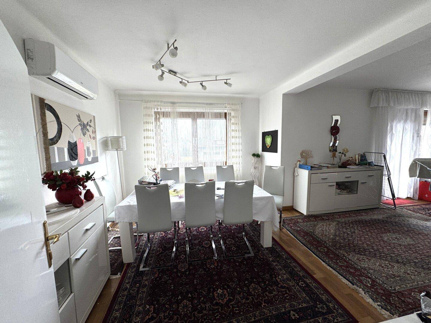 Helles Esszimmer mit großem Esstisch, weißen Stühlen und Klimaanlage.