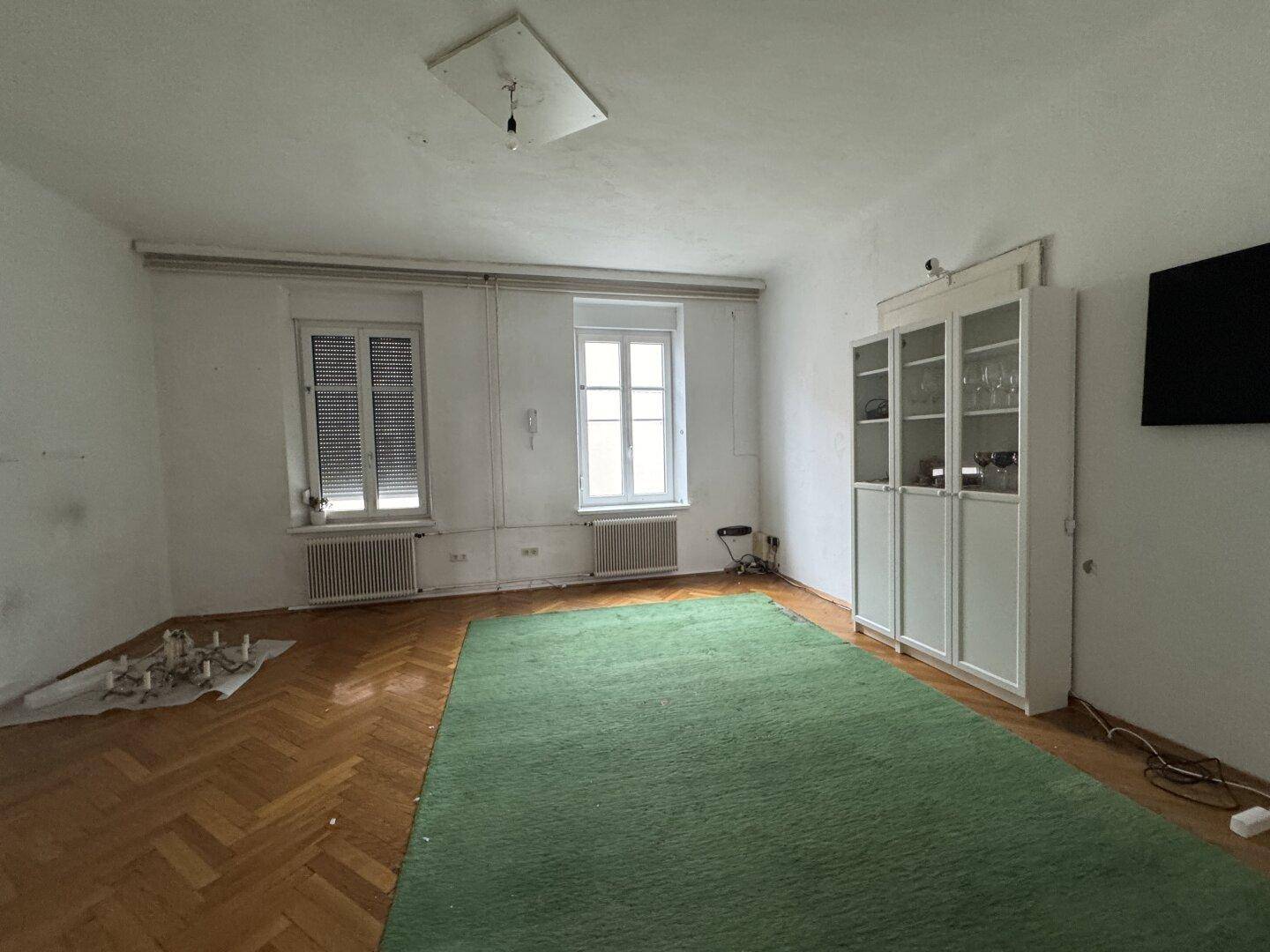 Geräumiges Wohnzimmer mit Parkettboden, grünem Teppich und Fenstern.