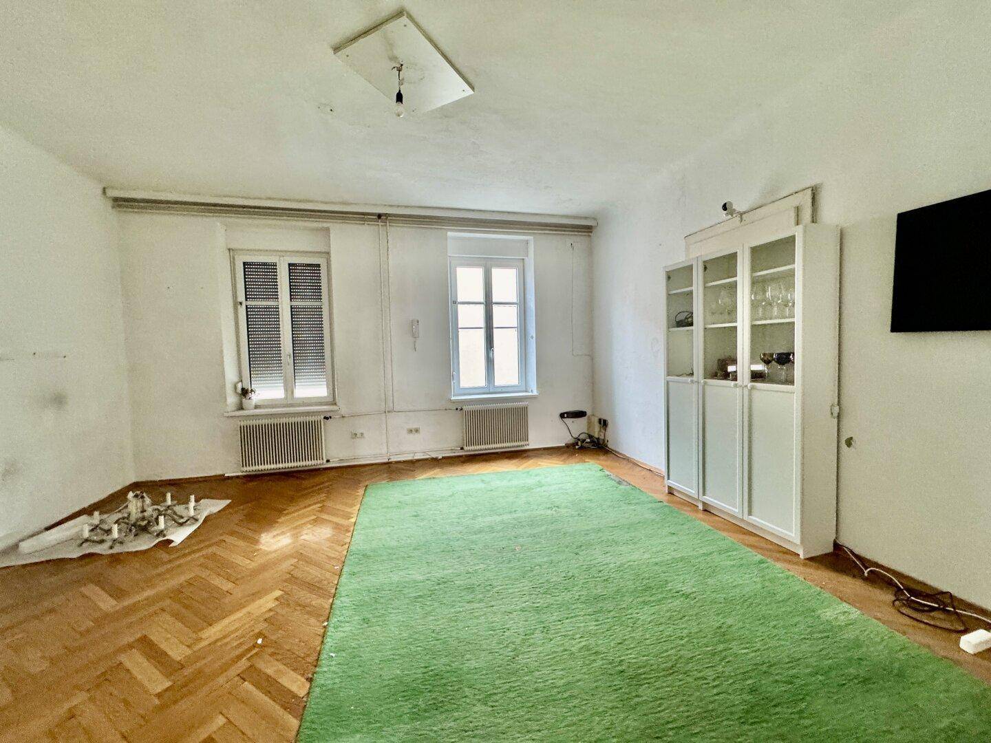 Geräumiges Wohnzimmer mit Parkettboden, grünem Teppich und Fenstern.