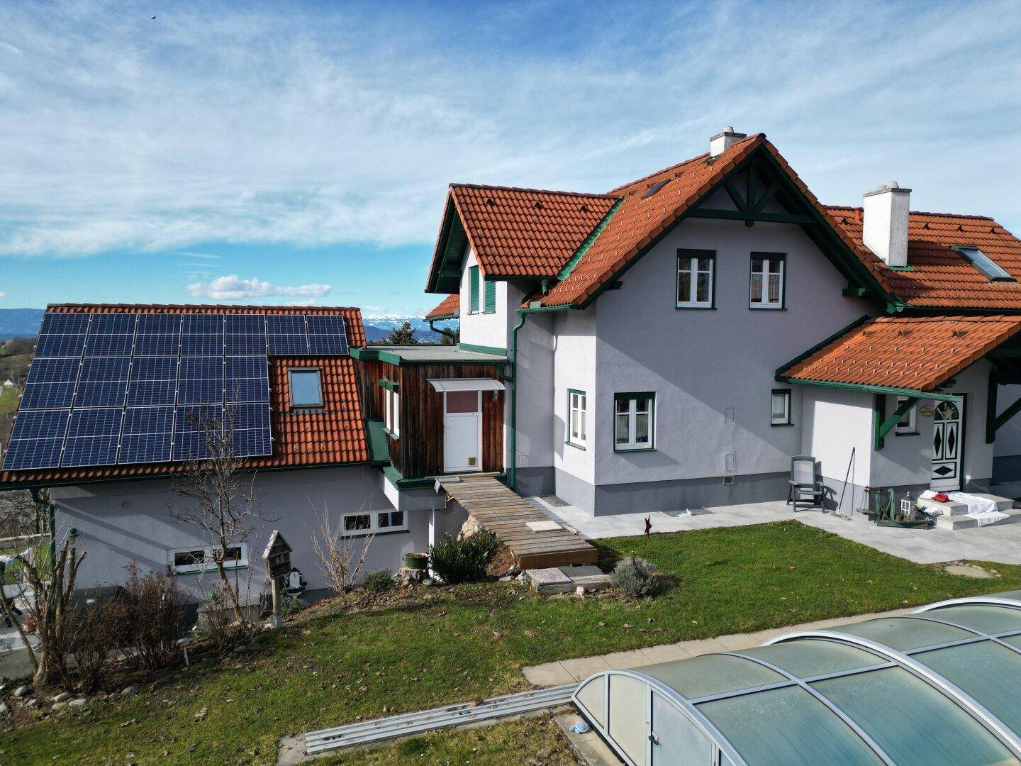 Haus mit Solaranlage, grünem Garten und überdachtem Pool unter strahlend blauem Himmel.
