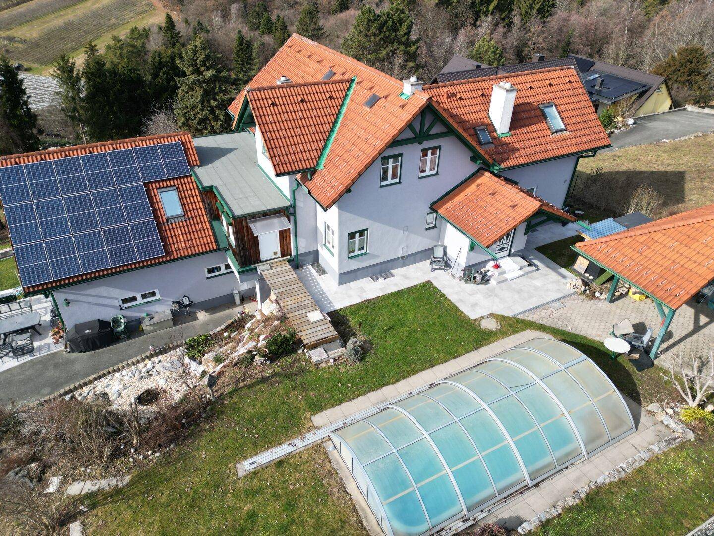 Luftaufnahme eines Hauses mit Solaranlage, Garten, überdachtem Pool und umliegender Naturlandschaft.