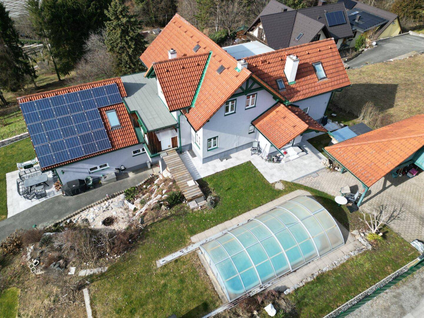 Luftaufnahme eines Hauses mit Solaranlage, Garten, überdachtem Pool und umliegender Naturlandschaft.