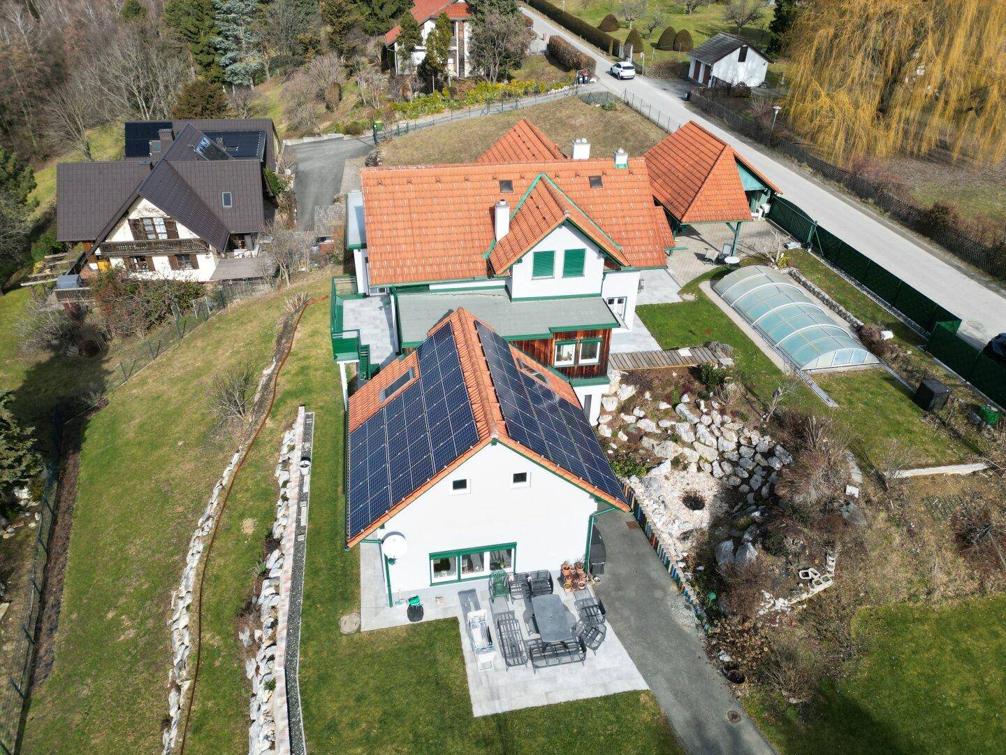 Luftaufnahme eines Hauses mit Solaranlage, Garten, Pool und umliegender Bebauung und Straße.