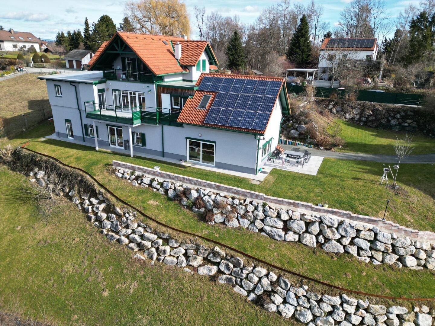 Luftaufnahme eines Hauses mit Photovoltaikanlage, Steinterrasse und weitläufigem Rasen.