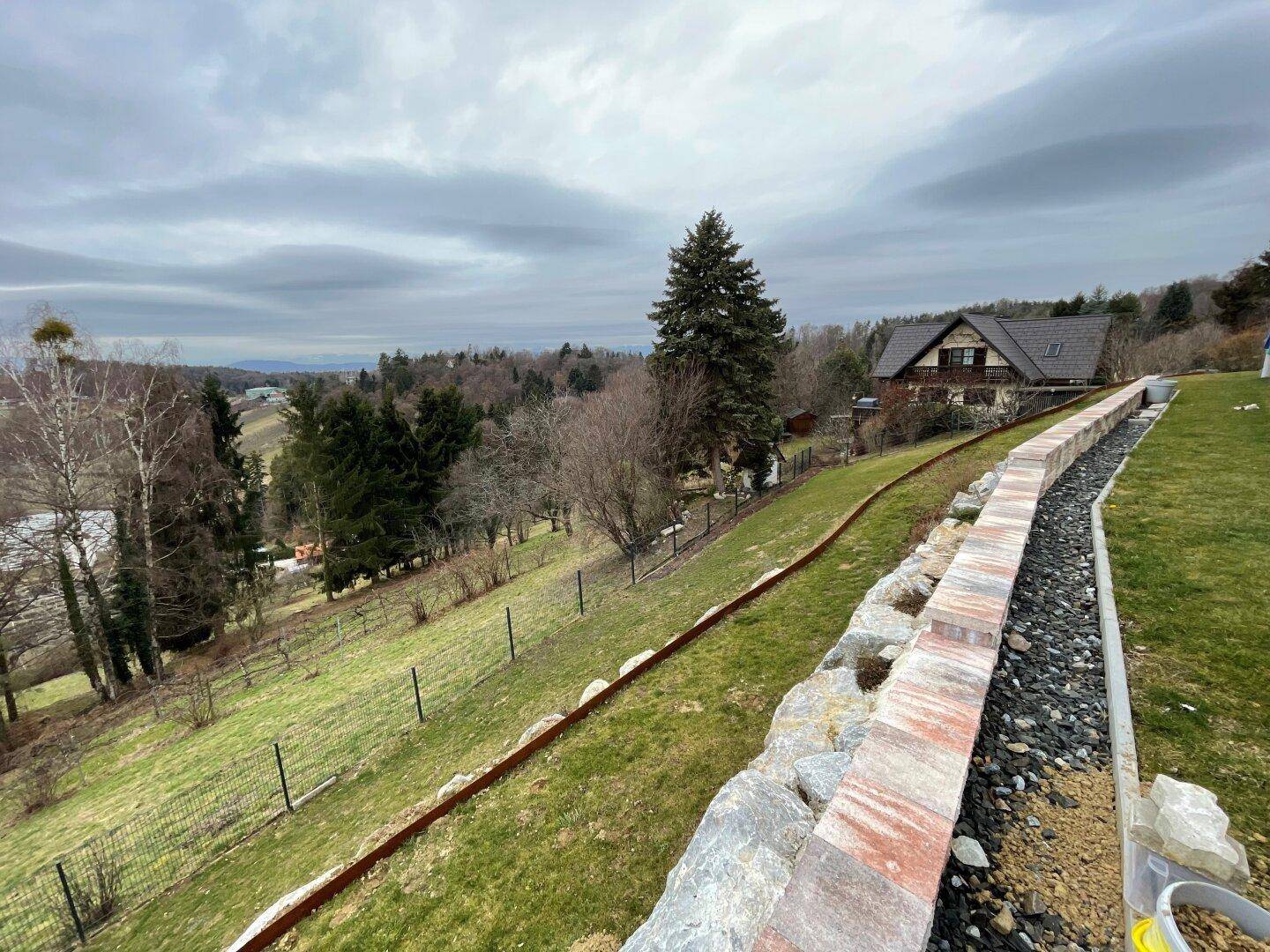 Hanglage mit terrassiertem Gartenbereich, Steinmauer und weitem Blick in die grüne Landschaft.