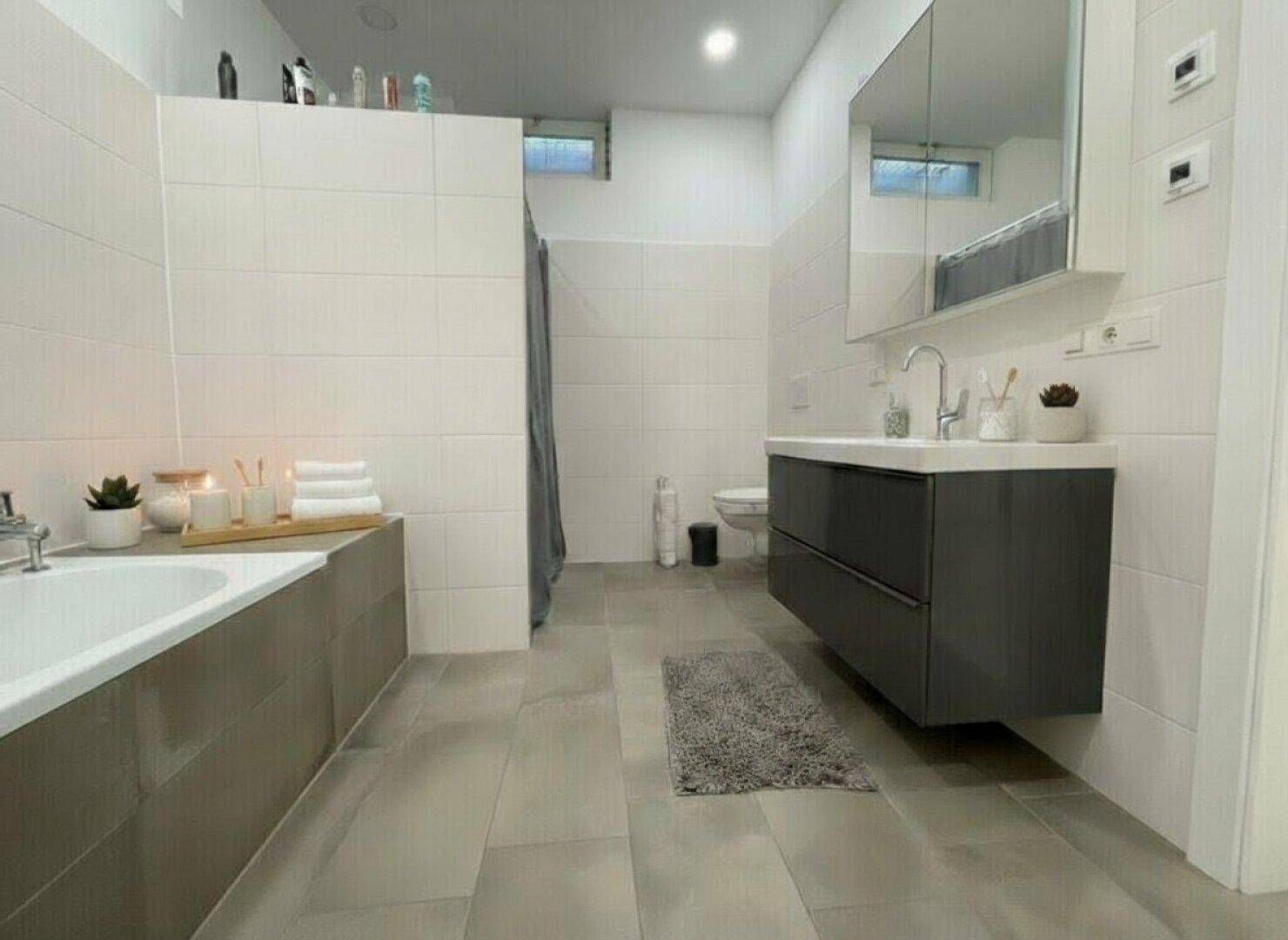 Modernes Badezimmer mit Badewanne, schwebendem Waschtisch und großen Fliesen in neutralen Farben.