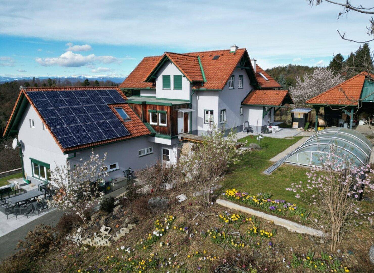 Haus mit Solaranlage, blühendem Garten, Pool und Bergblick unter strahlend blauem Himmel.