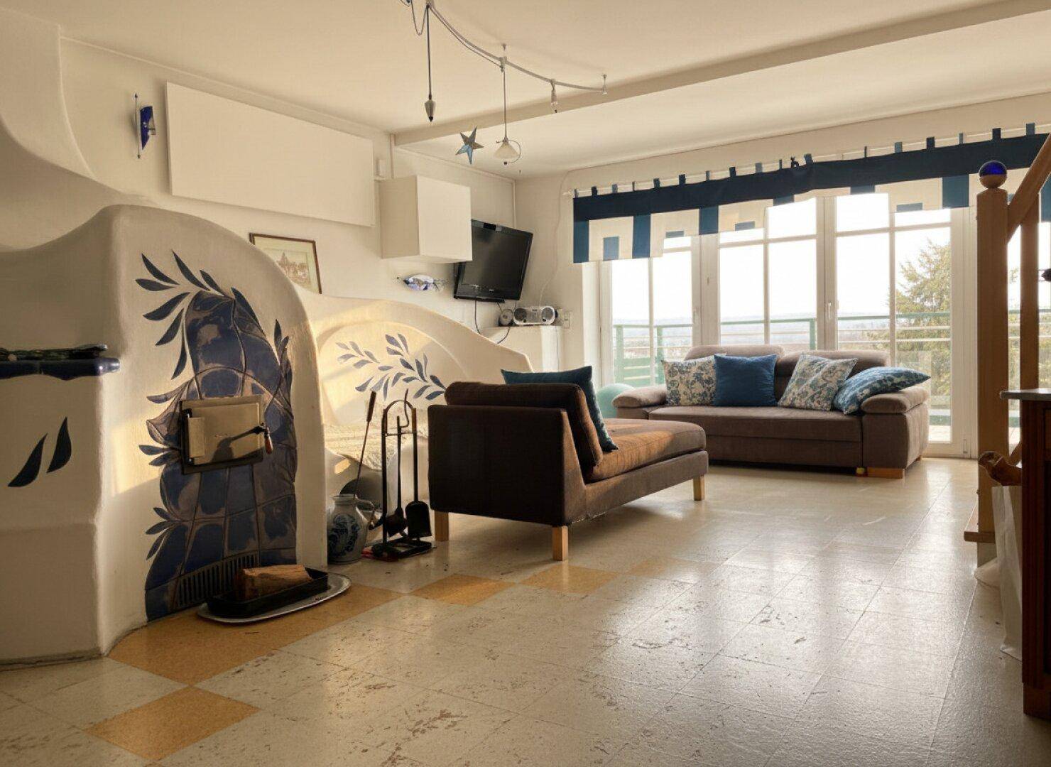 Helles Wohnzimmer mit traditionellem Kachelofen, bequemen Sofas und großen Fenstern.