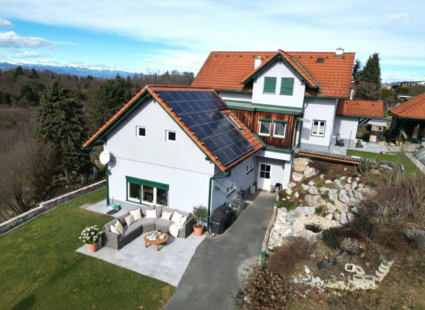 Haus mit Solaranlage, großer Terrasse, bequemen Gartenmöbeln und Bergblick unter blauem Himmel.