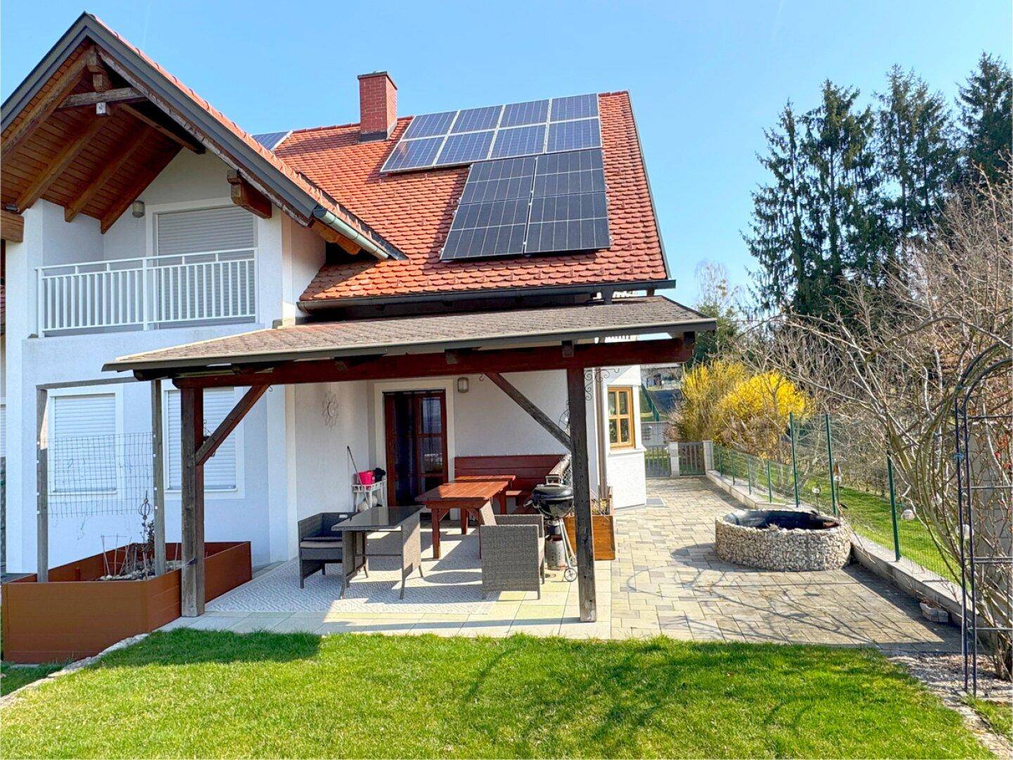 Detailansicht der überdachten Terrasse mit Esstisch, Grill und angrenzendem Feuerplatz im Garten.